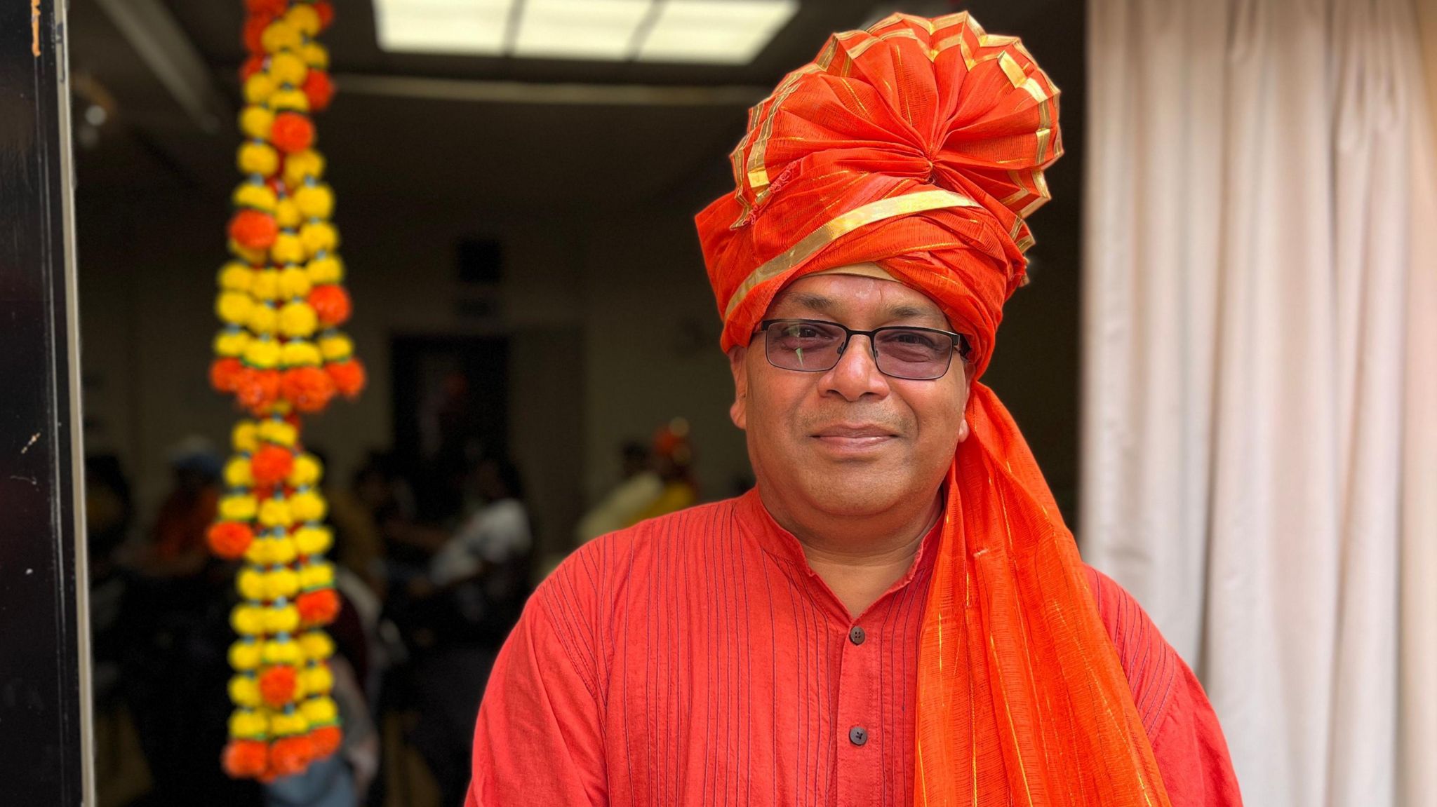 A man wears an orange headdress with a gold trim and a pleated fan effect on top