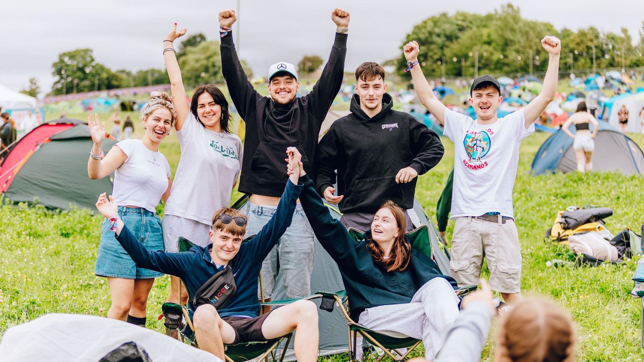 Campers in the campsite cheer, with their arms aloft. Two at the front are sat on camp chairs, the five behind are standing.