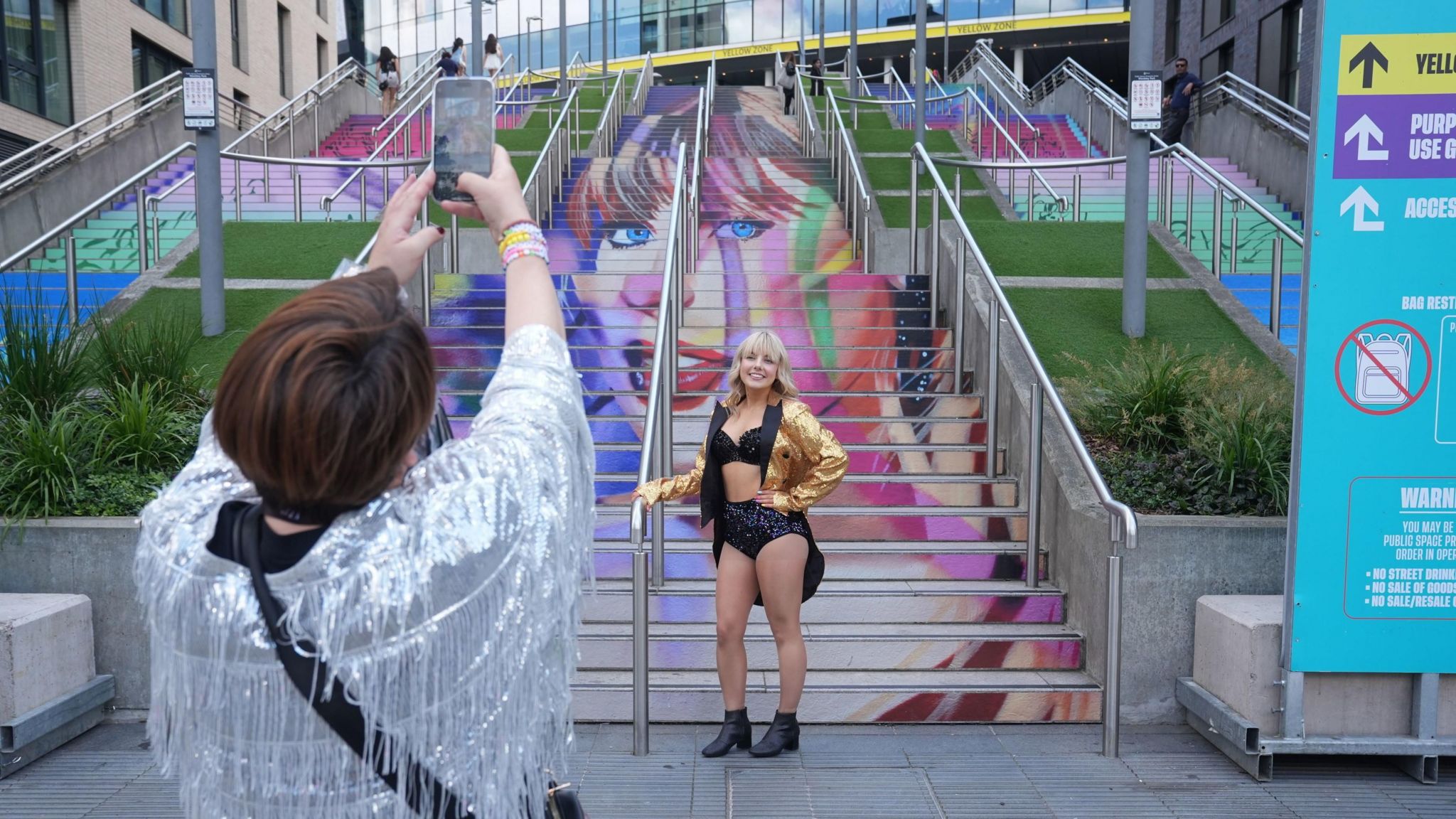 A blonde woman in a gold jacket, black sparkly hot-pants and a black sparkly crop top, having her photo taken by a woman in a silver sparkly jacket in front of stairs that have Taylor Swift's face painted on them in multi colours