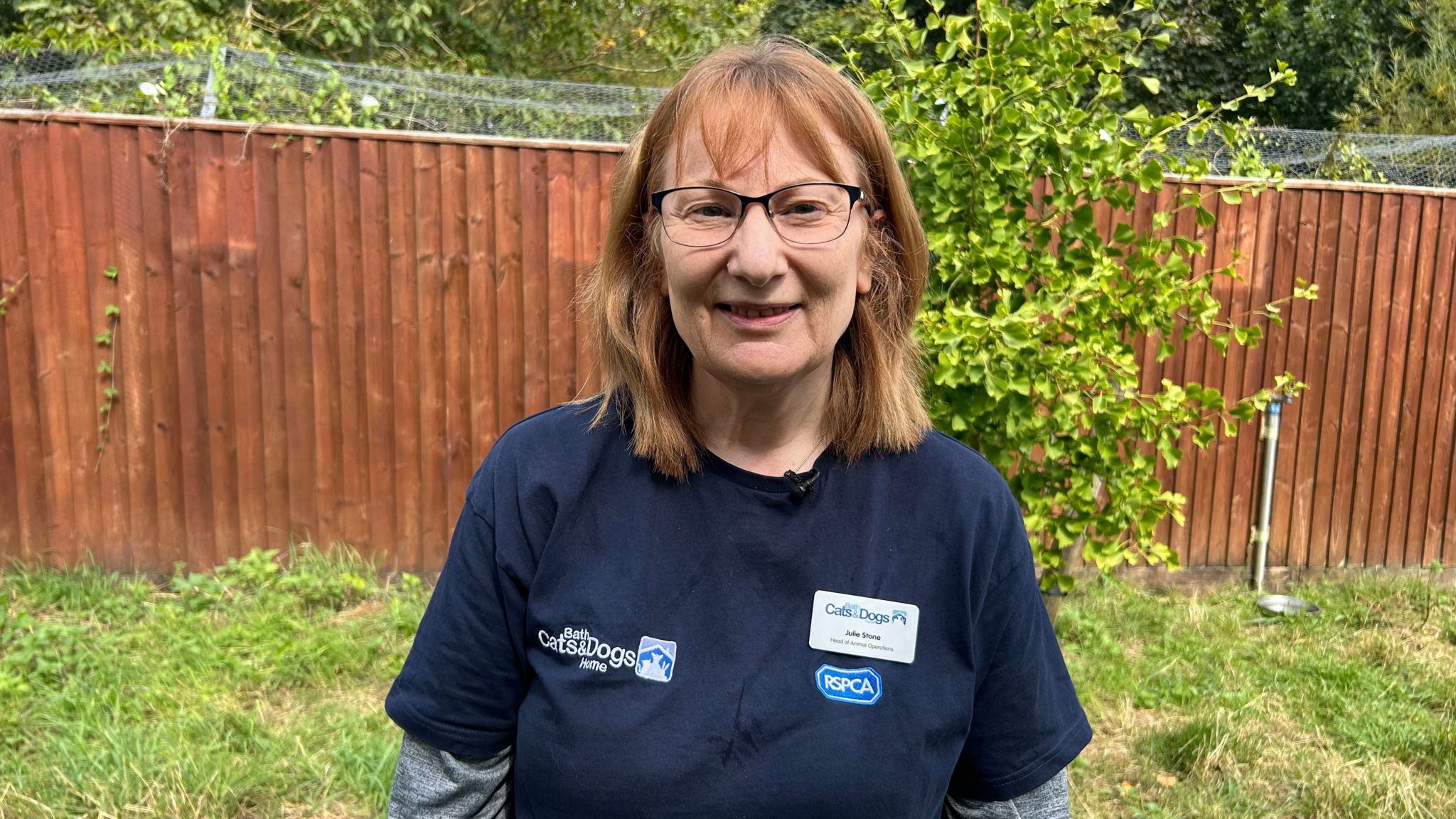 Photo of Julie Stone, head of animal operations at Bath Cats and Dogs Home. She is wearing a blue t-shirt and glasses, and has shoulder length strawberry blonde hair. She is standing on grass in front of a wooden fence.