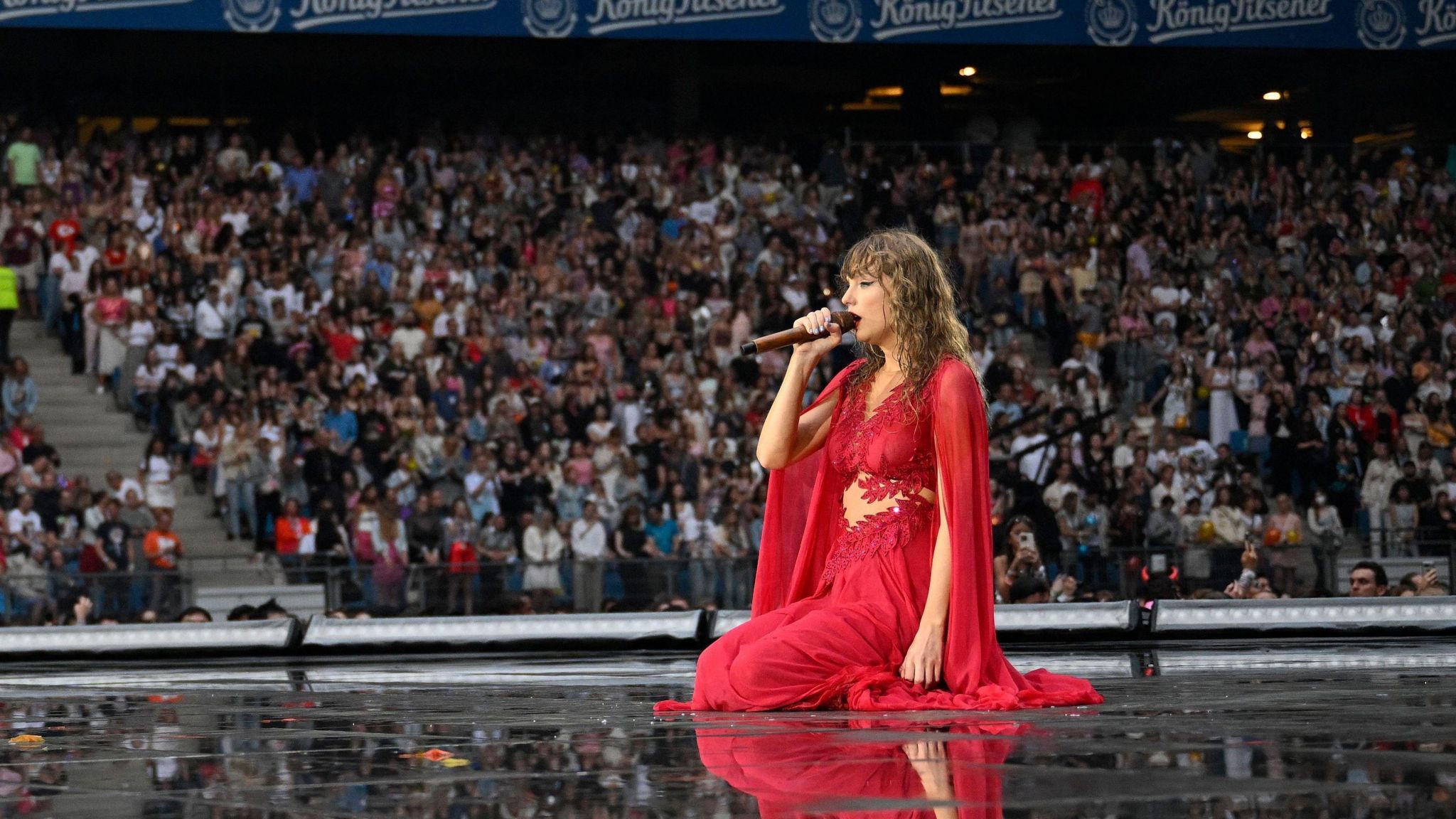 Taylor Swift performing in Hamburg as part of her Eras tour. Taylor, dressed in a flowing red gown, kneels on stage, holding the microphone towards her. Crowds can be seen in the stadium behind her. 