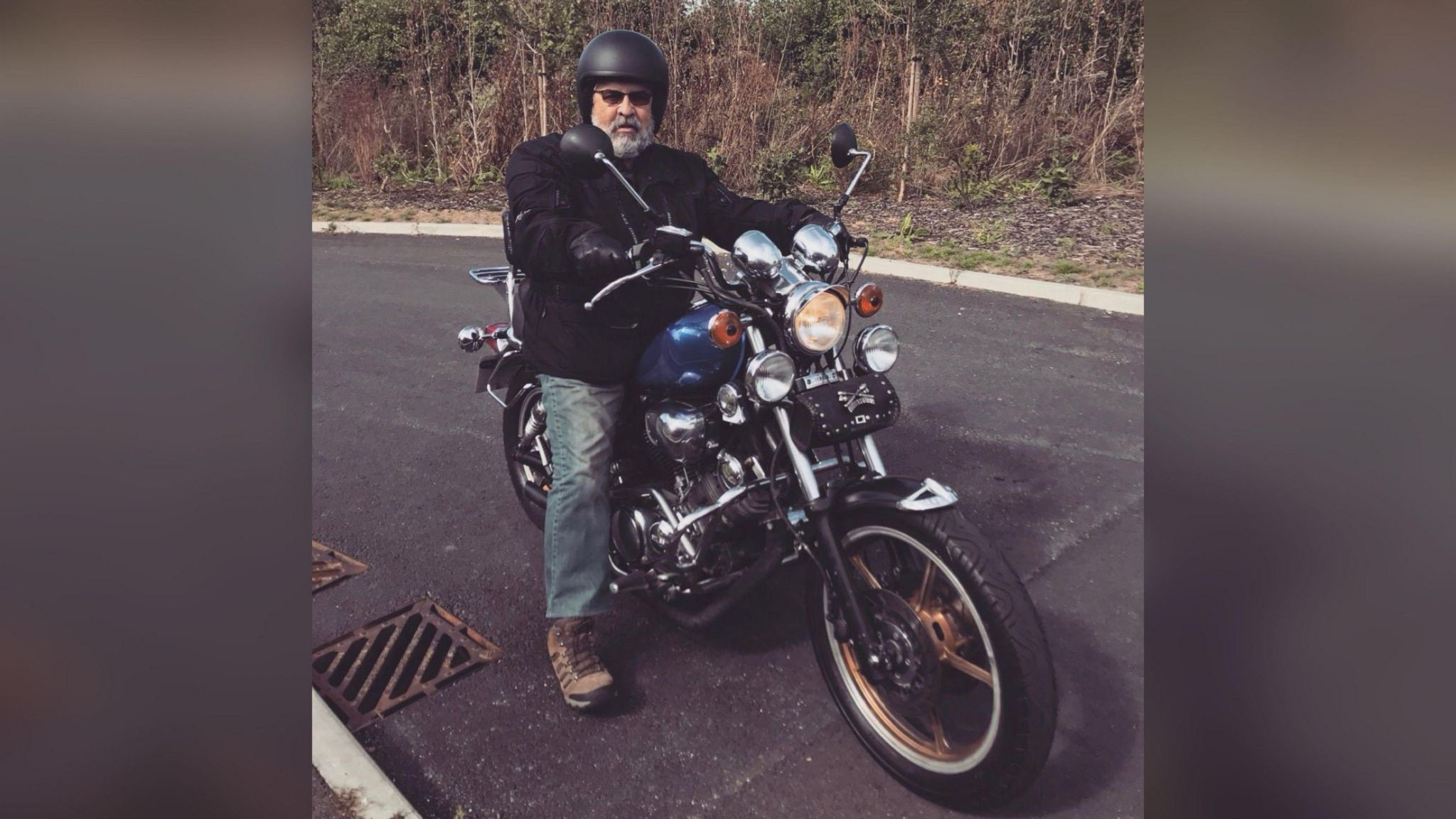 Steve Macdonald astride a black motorbike wearing light jeans, a dark motorbike jacket, black helmet and sunglasses.