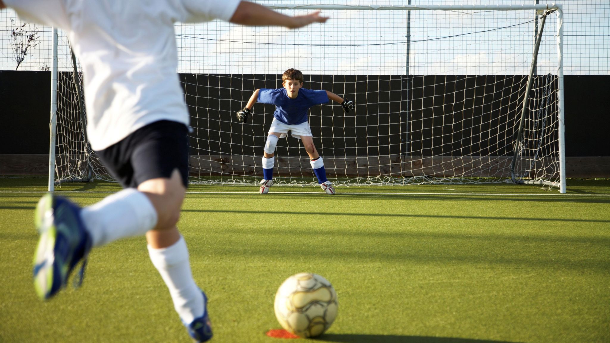 Kid kicking a football at a goalkeeper in a goal