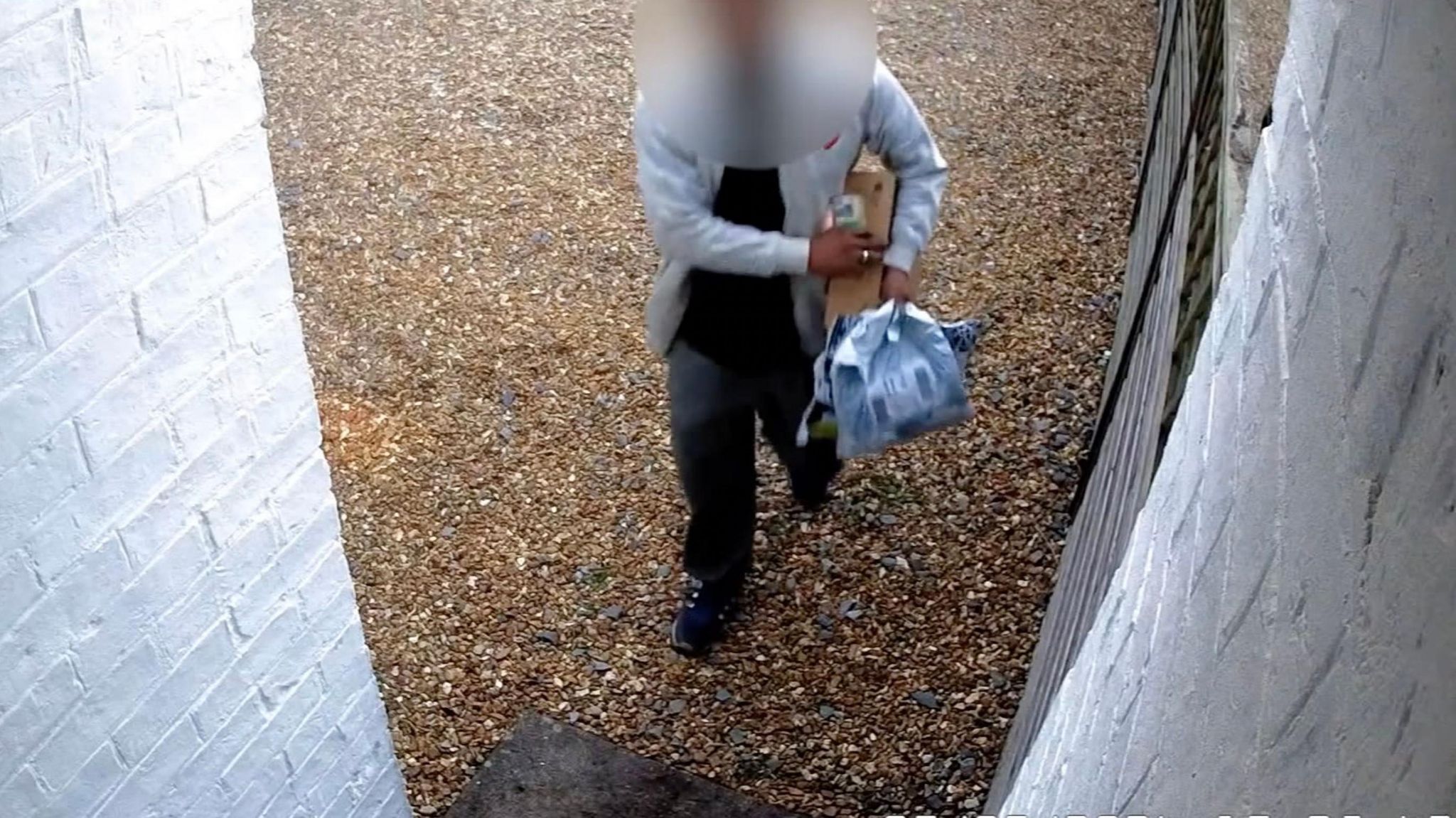 A security camera image of a driver delivering parcels to a house. They are wearing dark grey trousers and top and a light grey jacket and their face has been blurred.