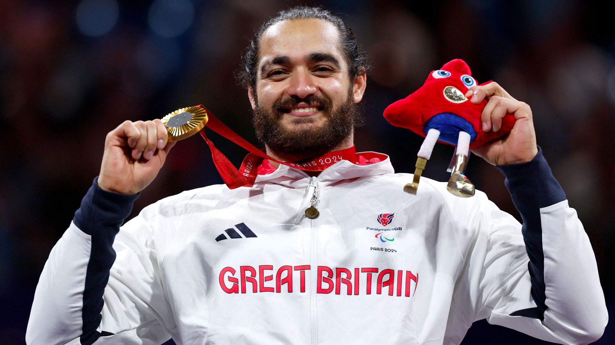 Dimitri Coutya. He has dark hair and a beard. He is pictured wearing a white jacket with Great Britain printed across the chest in red letters. He is holding a gold medal and a red Paralympic mascot soft toy. 