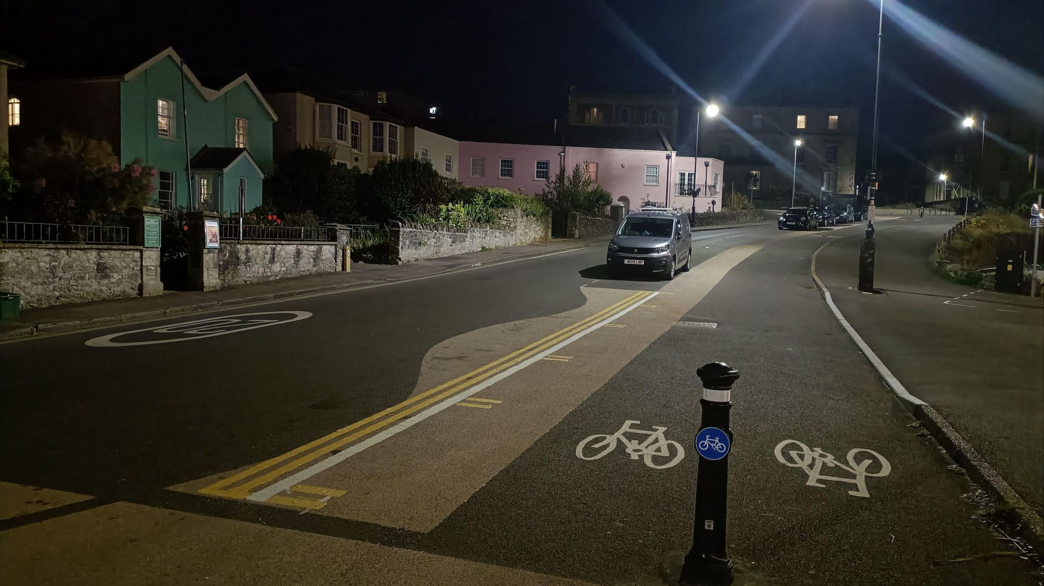 Cycle lane will bollard, next to straight double yellow lines, with wavy road markings, then the road running parallel to the left.