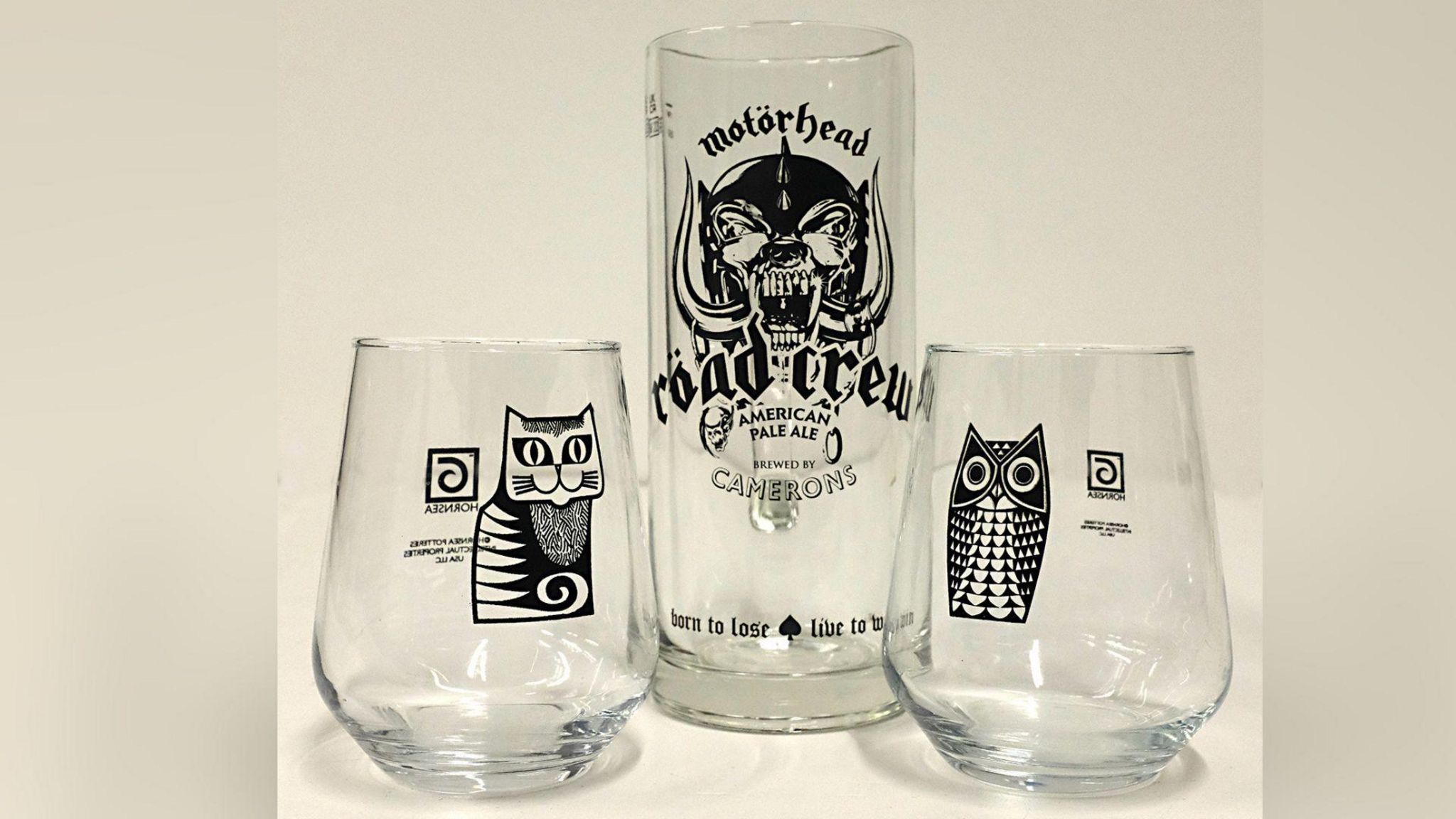 Three ale glasses on a shelf. One has a cat image, another an owl and a tall one in the centre has the words Motorhead and road crew.