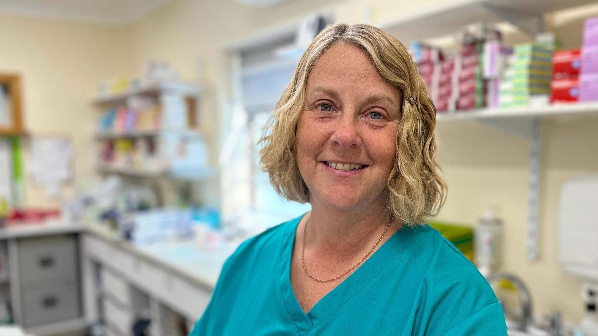 Denise Burton, who has blonde hair and wears green scrubs, stands in the dispensary in front of shelves of medication.