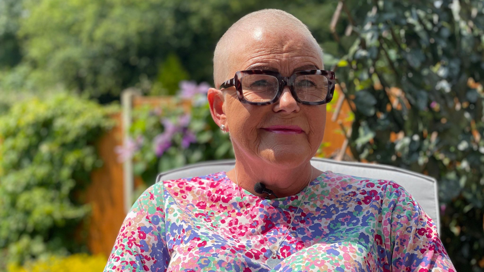 Tracie Mills in her garden. She has shaved hair and wears glasses with a slight smile at the camera. She is wearing a floral dress