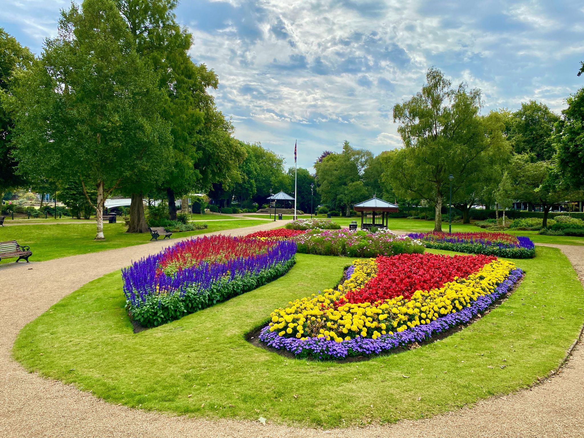 An oval area of grass with four floral beds around a circular central patch. They feature red blue and yellow flowers in varying arrangements, with pink and white in the central bed.