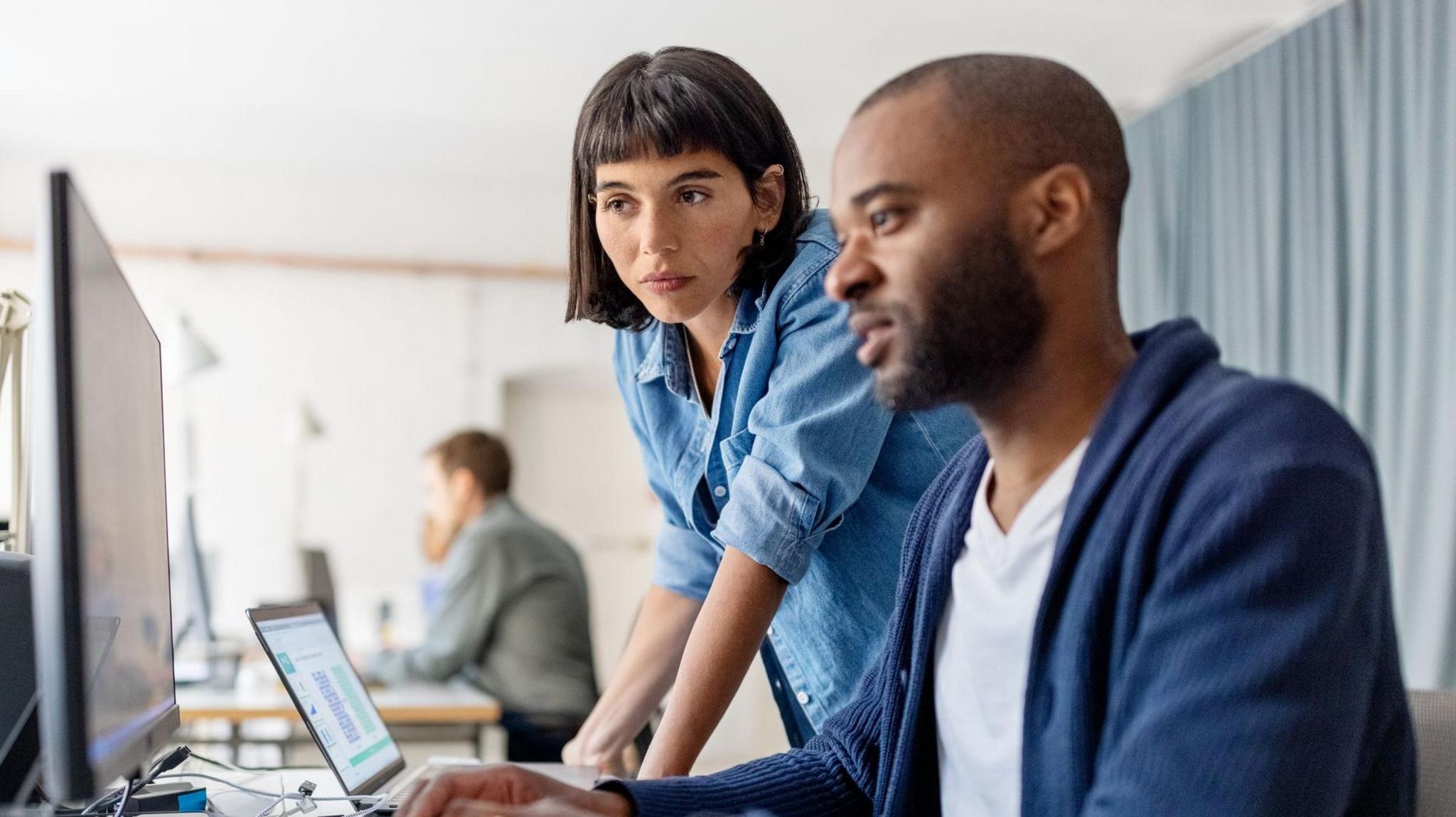 Woman leaning on man's desk as they work together on something on a computer screen