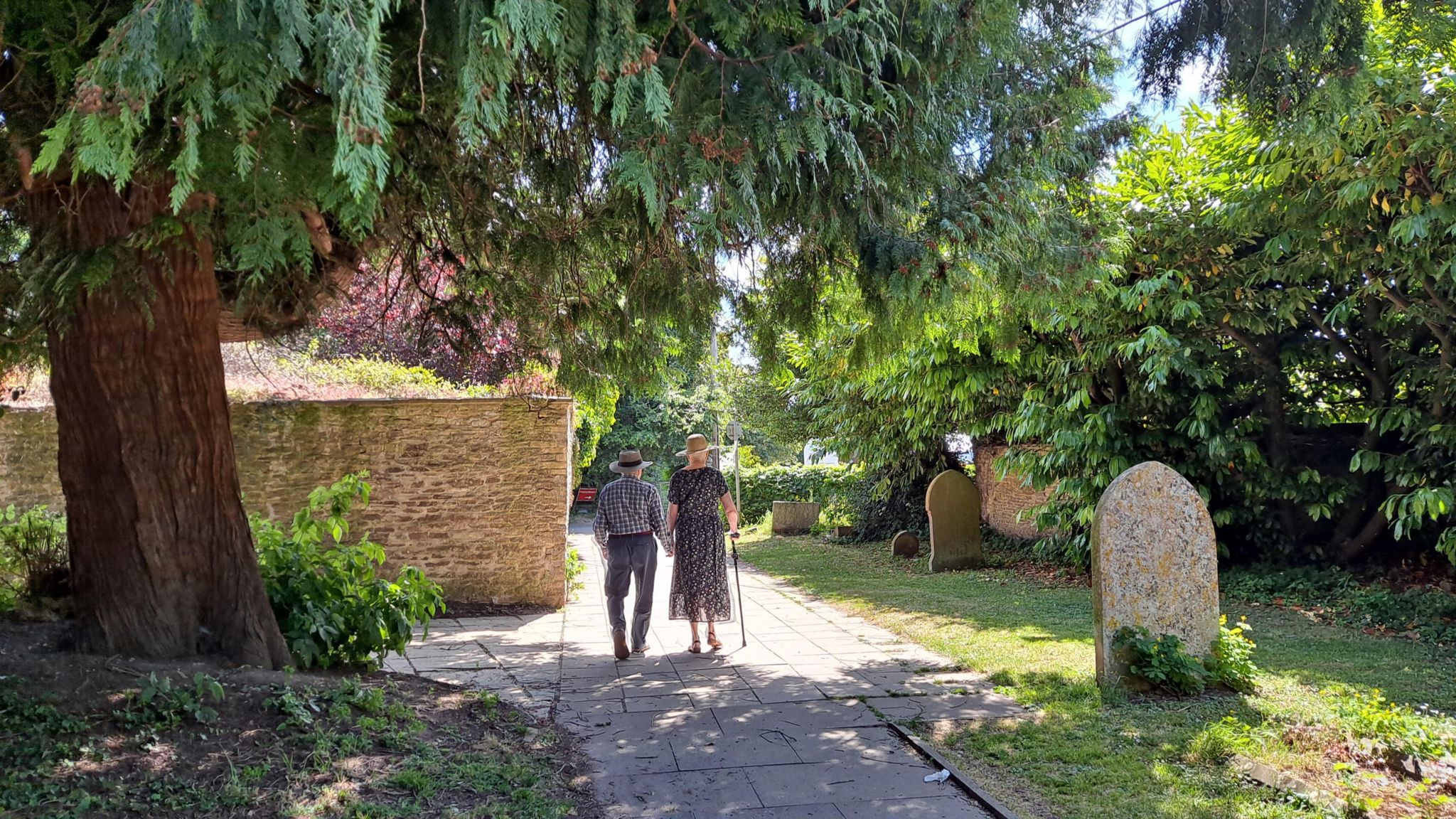 WEDNESDAY - A man and woman holding hands as they walk through a graveyard on a sunny day. They are both wearing straw hats. The woman is using a walking stick and is wearing a dress, the man is wearing a shirt and trousers. They are walking under a pine tree into the sunshine. On their left is a brick wall, on the right are several gravestones and a tall green hedge