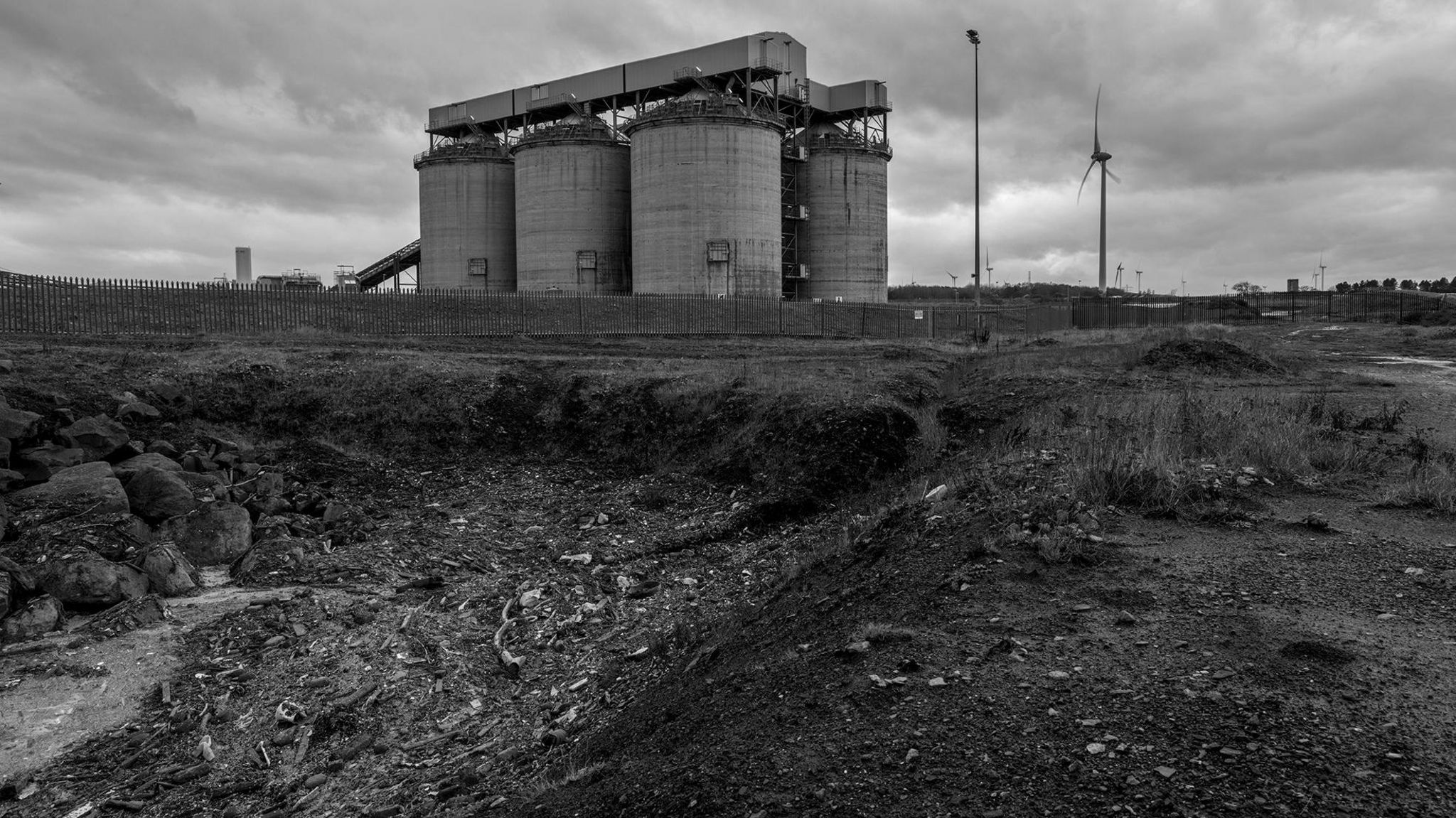 A black and white shot of an beach full of debris in the foreground with the chimneys of a power station in the background 