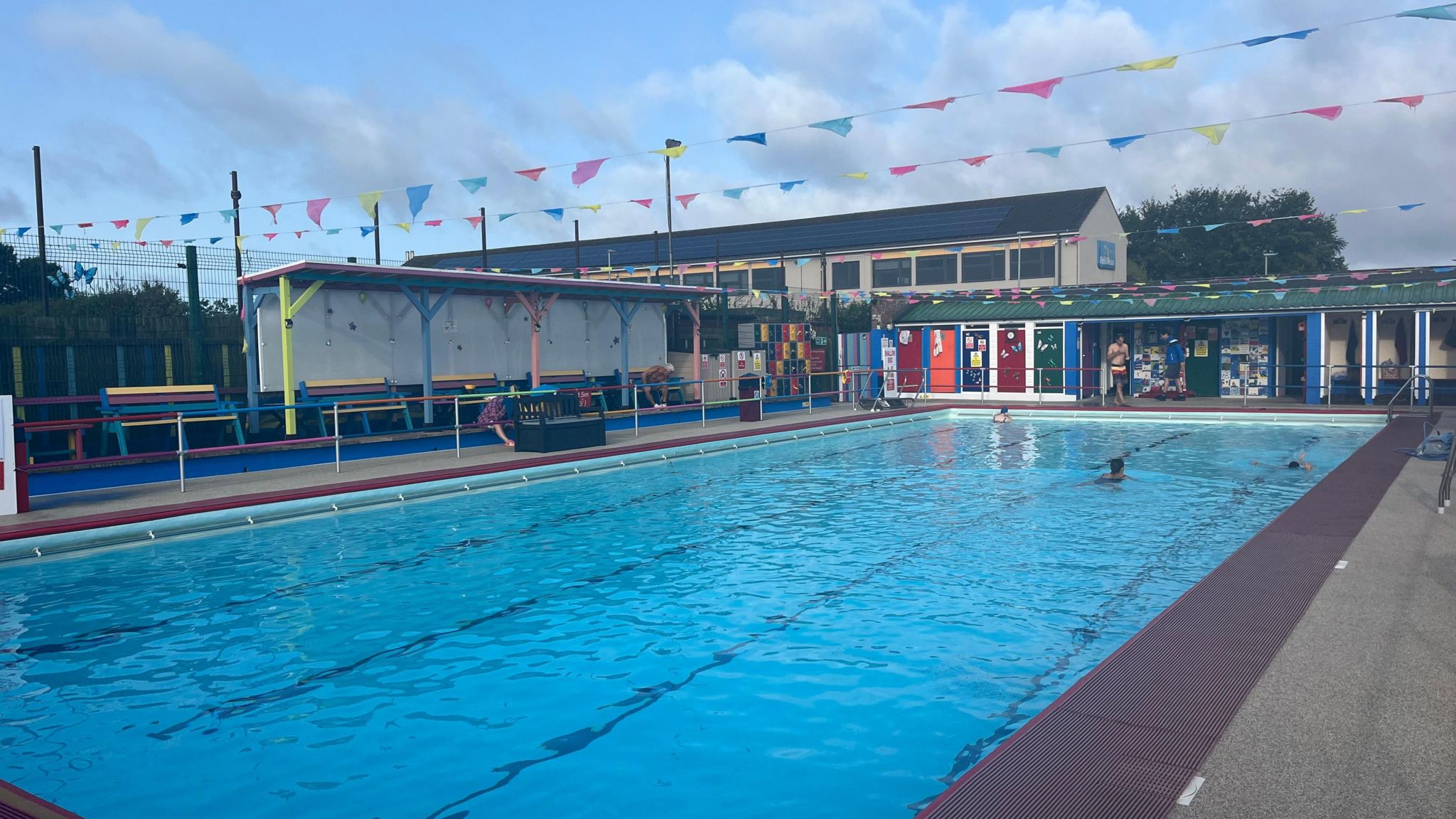 An outdoor pool with colourful changing rooms around it, benches and bunting above