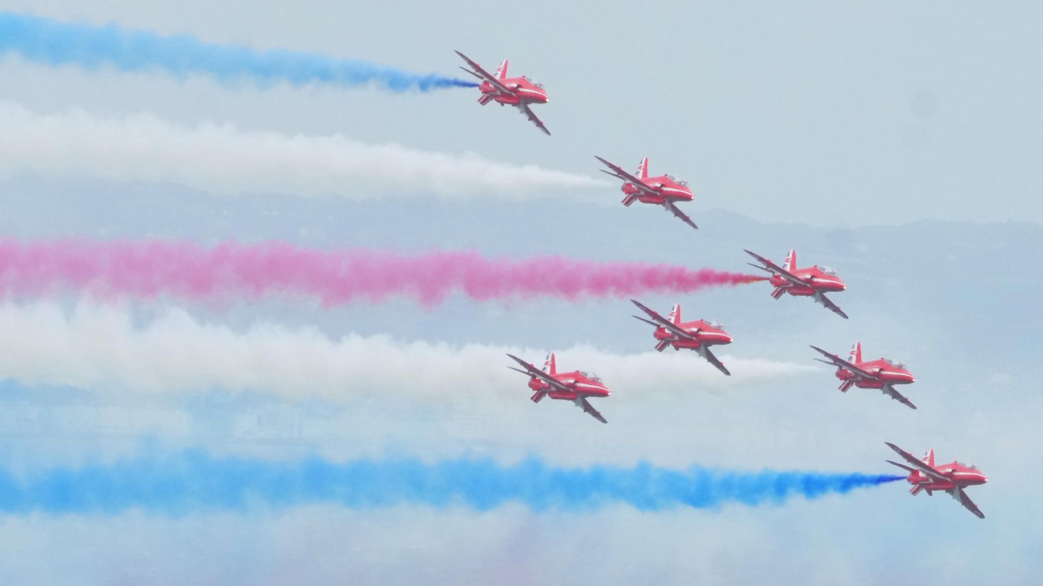 Seven bright red planes fly in the sky with blue, white and red vapour trails. 