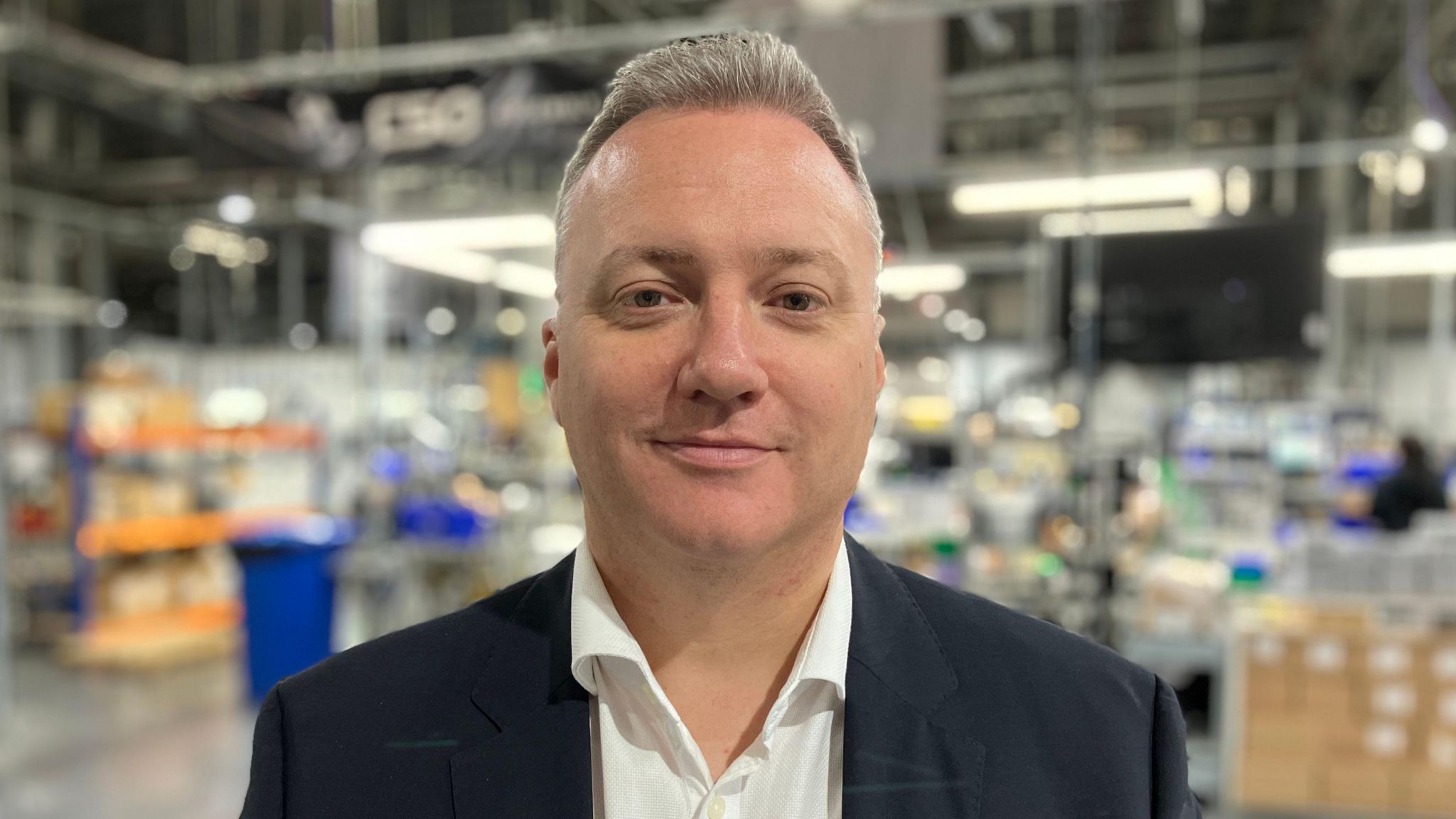 Steve Elwell looking into camera wearing suit jacket and white shirt while standing on factory floor