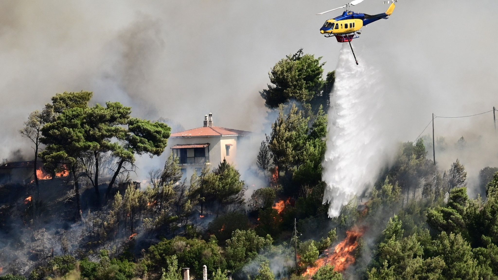 A helicopter flies, dropping water over a fire burning near a home