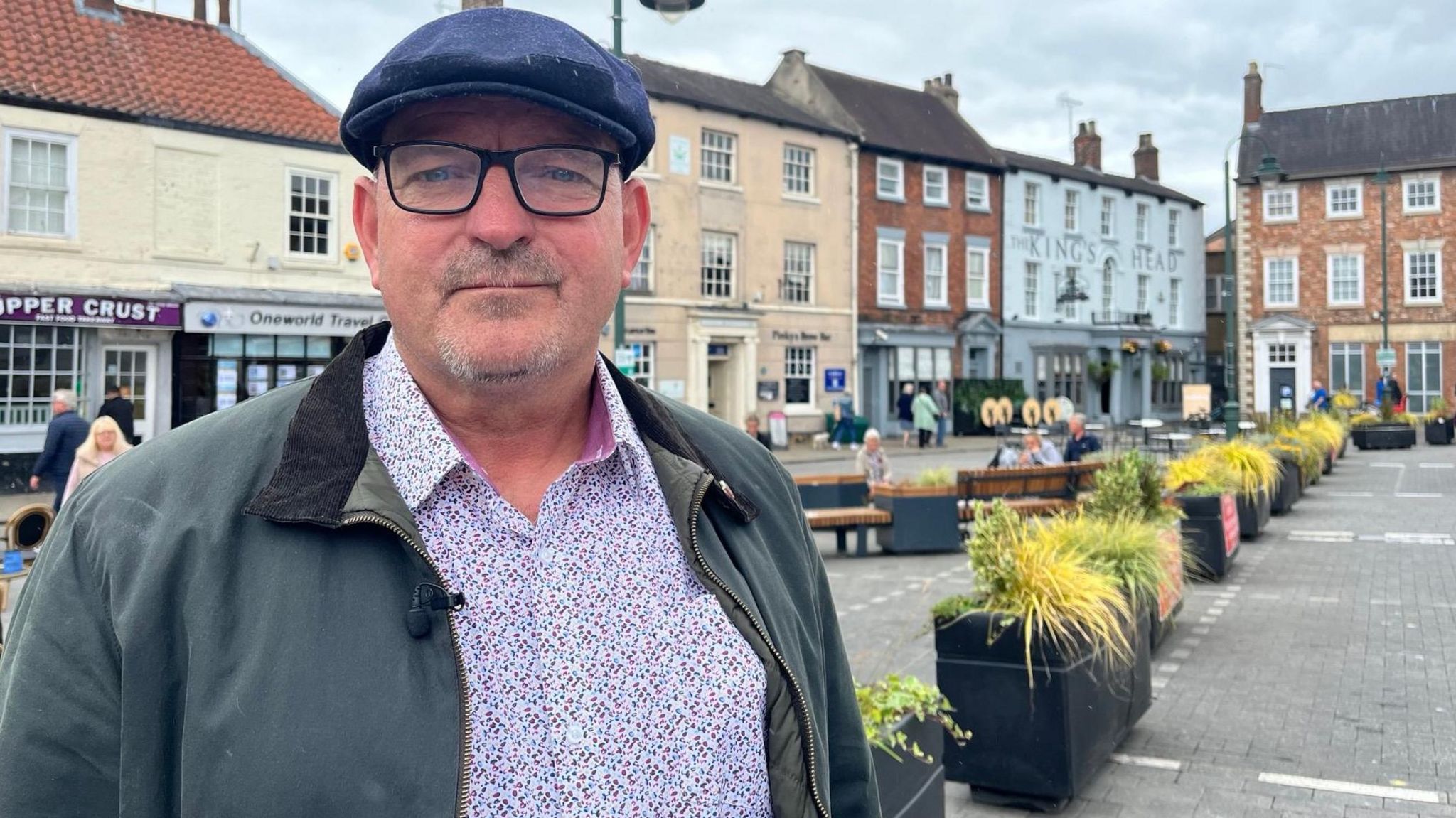 Kevin Smart is a middle-aged man with a goatee beard and moustache, wearing a flat cap and glasses, standing in front of a row of parking spaces in a market place wearing a cap, glasses and purple-speckled shirt