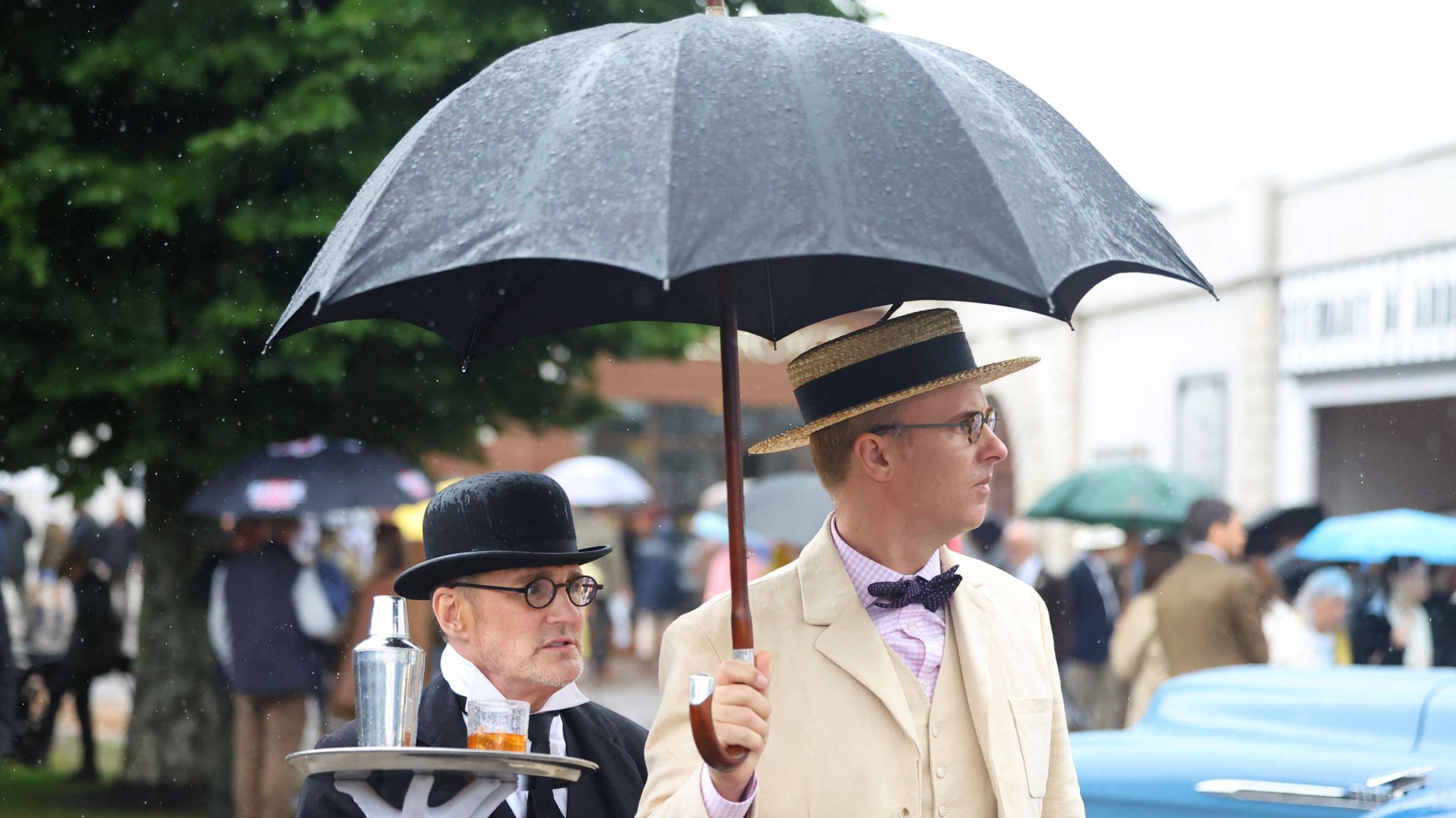 Two smartly dressed men shielding under an umbrella during a car-racing festival.