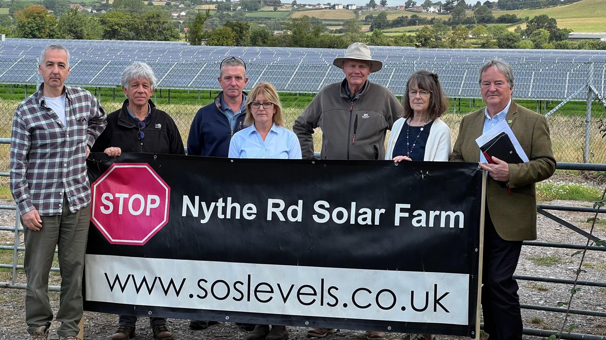 Campaigners stand with a banner in front of a field of solar panels at on the Somerset levels. It reads "Stop Nythe Rd Solar Farm"