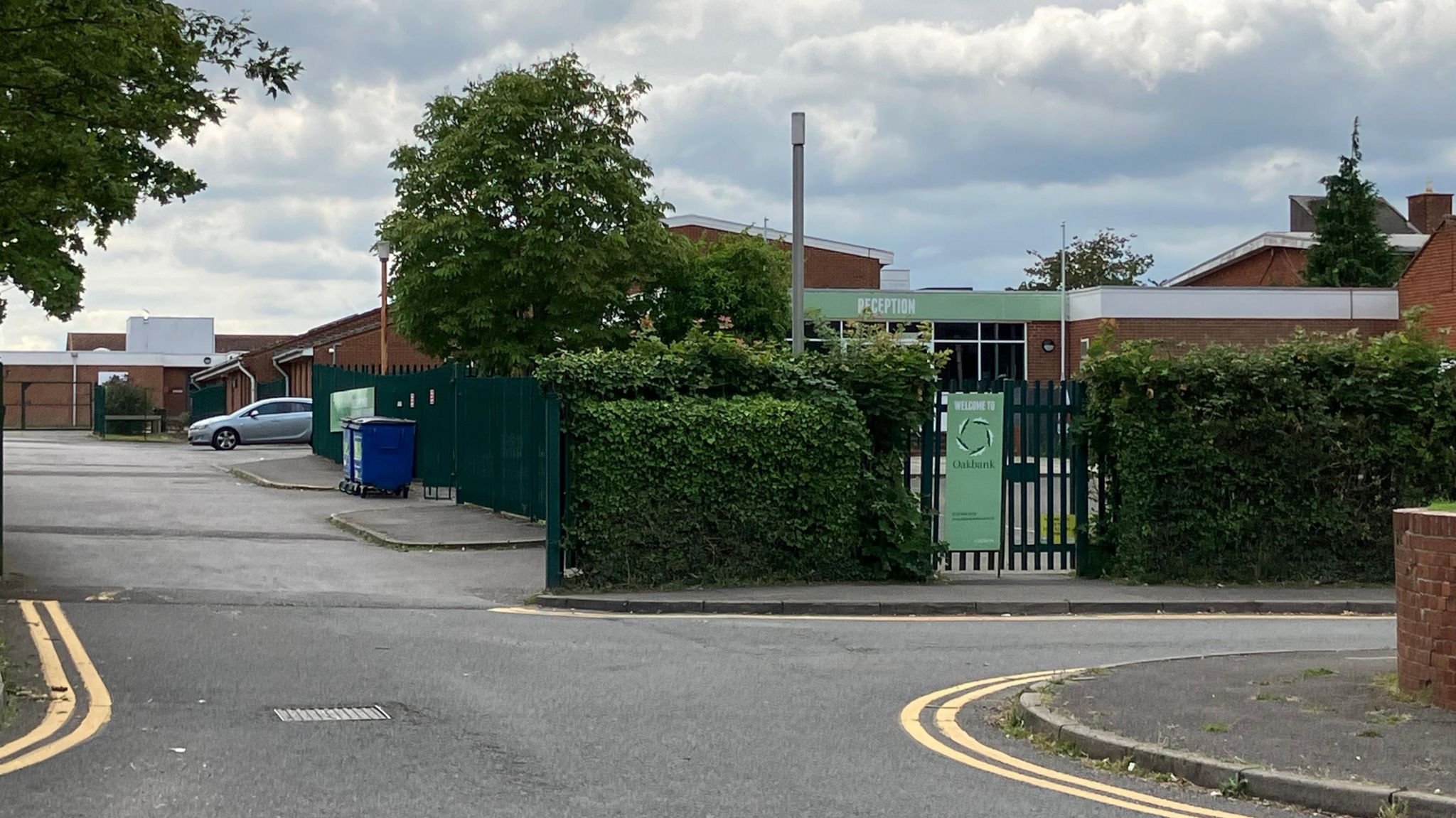 Oakbank Secondary School site, with a school gate, the school sign and a parked silver car in the distance