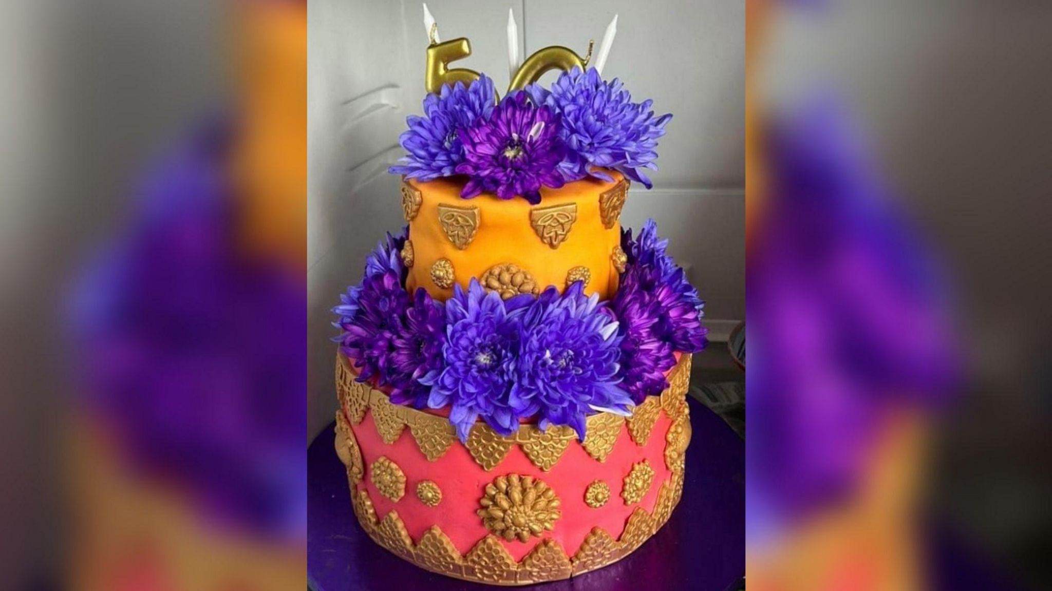 A two-tier cake decorated with gold icing. The first tier is red and the second is orange. The cake is topped with purple flowers.