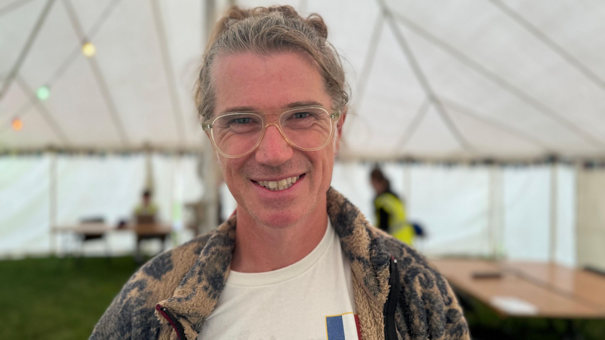 A man with grey/brown hair tied in a bun, smiles at the camera. He is wearing glasses and a brown jacket with grey patterns. He is standing inside a large white tent.