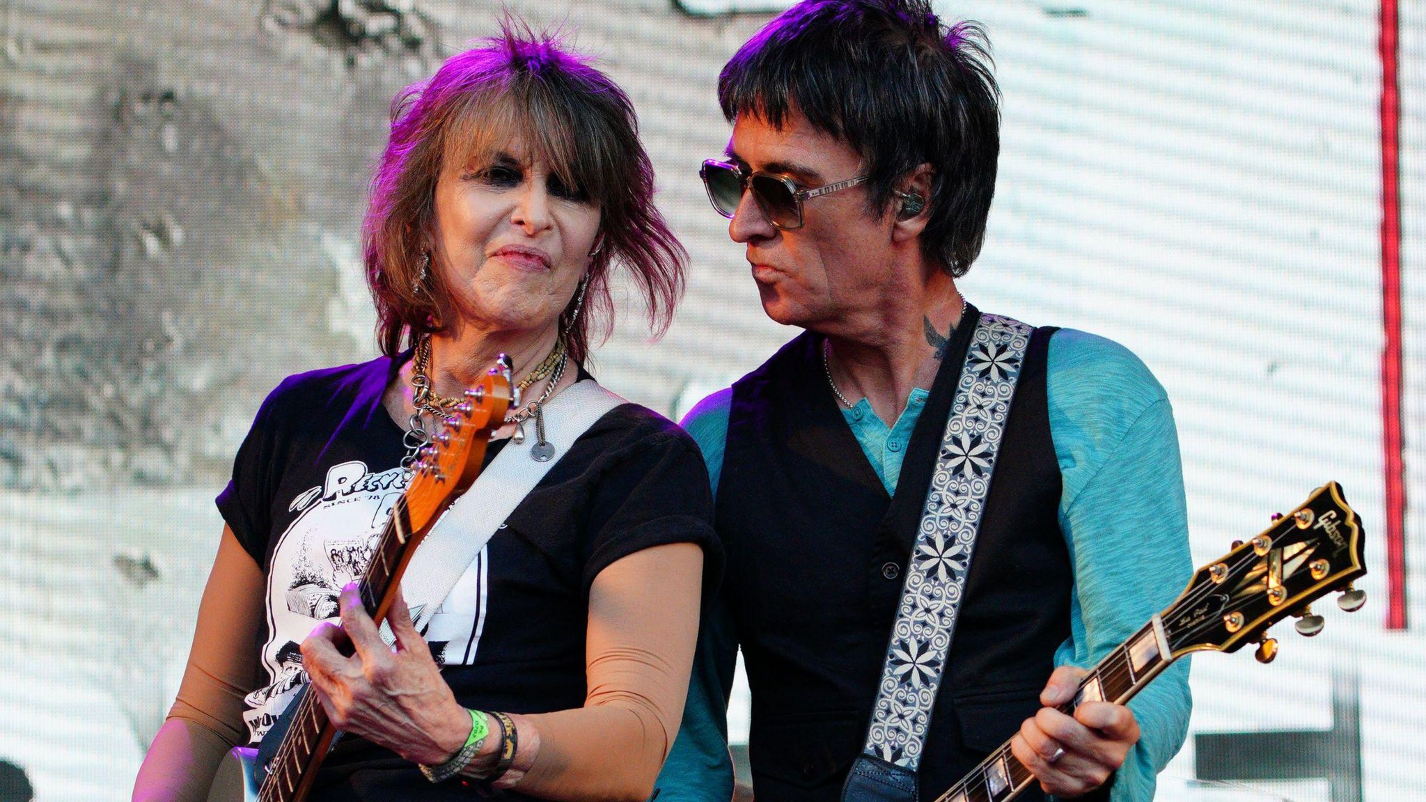 Chrissie Hynde from the Pretenders, wearing a black and white T-shirt and holding a guitar, with Johnny Marr, who wears sunglasses, a teal-coloured long-sleeved T-shirt and black waistcoat and holds a guitar, performing on stage at Glastonbury 