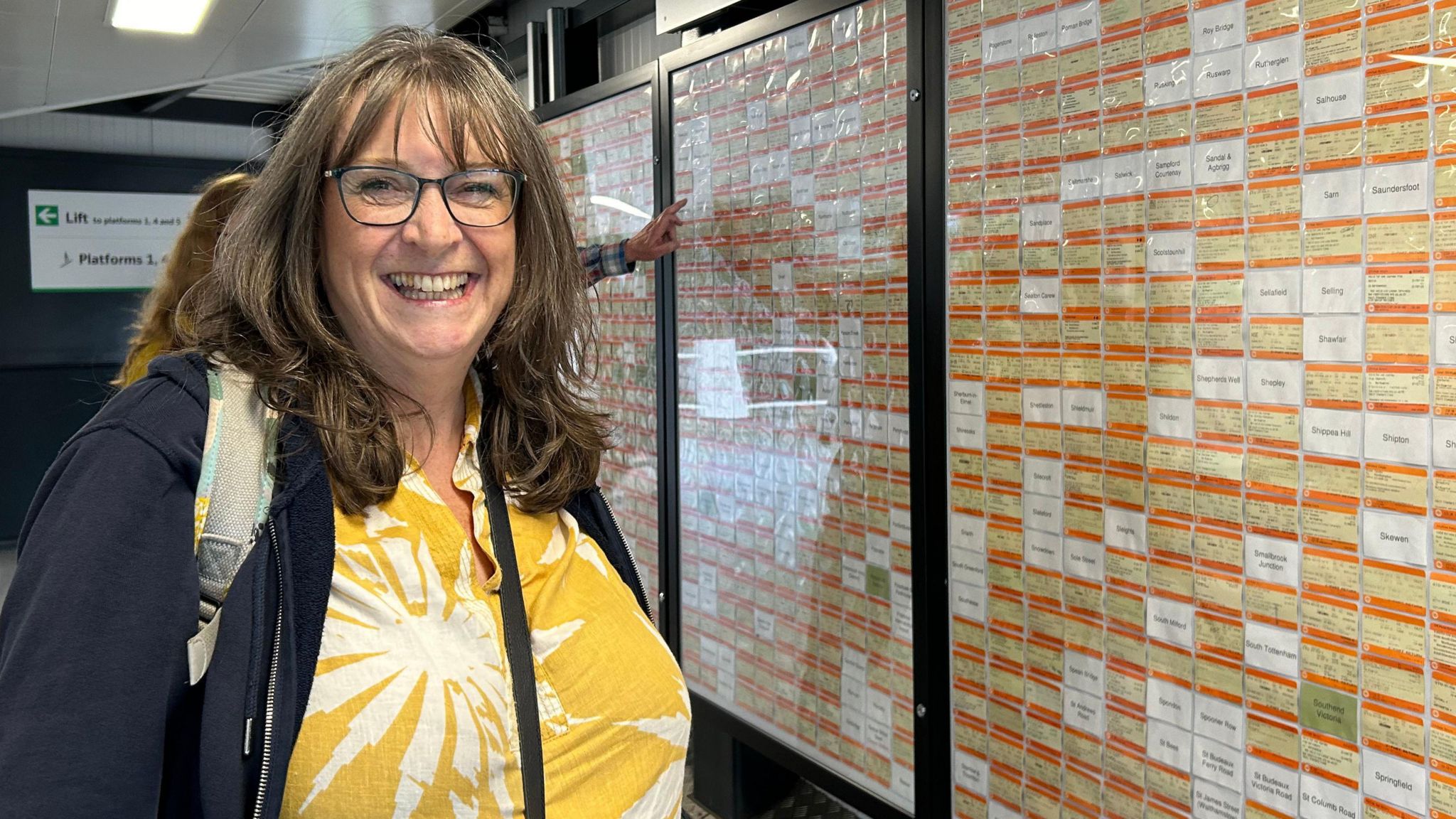 Carol Puckett with mid-length brown hair, wearing a yellow and white floral shirt and a dark zip-up sweatshirt. She is standing next to a display of bright orange train tickets