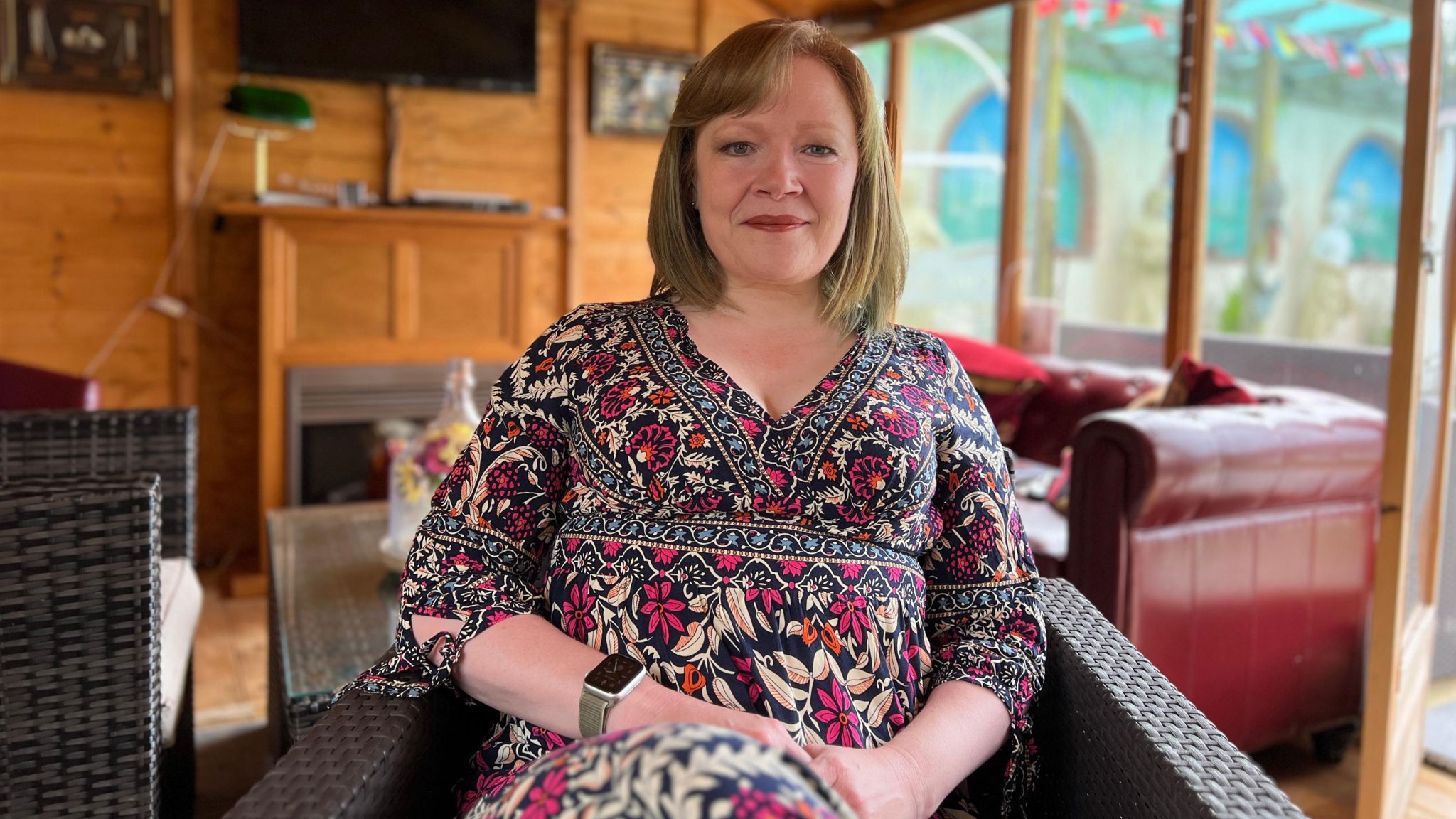 Lorna smiles at the camera as she sits on a chair with a sofa and a table behind her