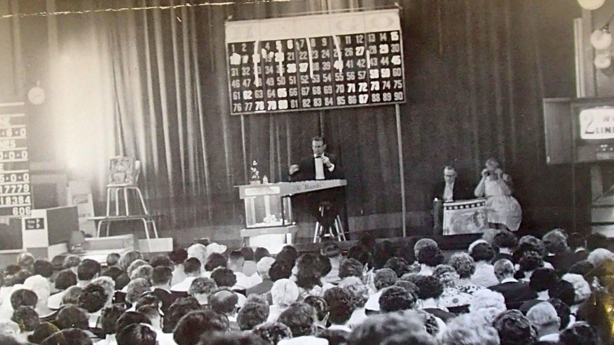 A crowd of people look at the stage where a man in black tie is sitting below a sign with numbers on them
