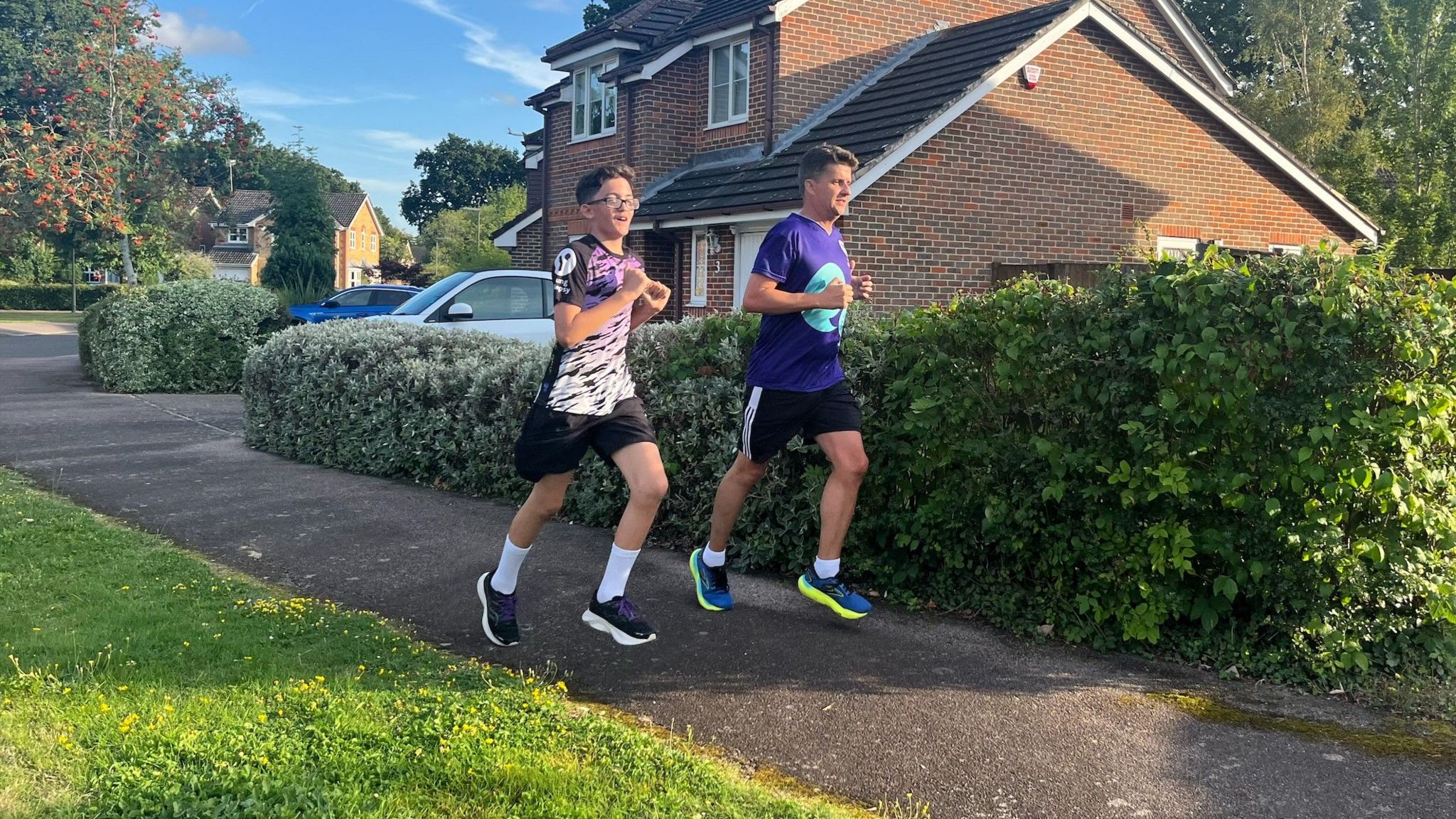 13 year old boy with brown hair and glasses, wearing a black, white and purple running top, black shorts and trainers running next to his dad who is wearing a purple Young Epilepsy t-shirt, black short and yellow and blue trainers. They are running past green hedges in front of a house and blue sky near Copthorne.