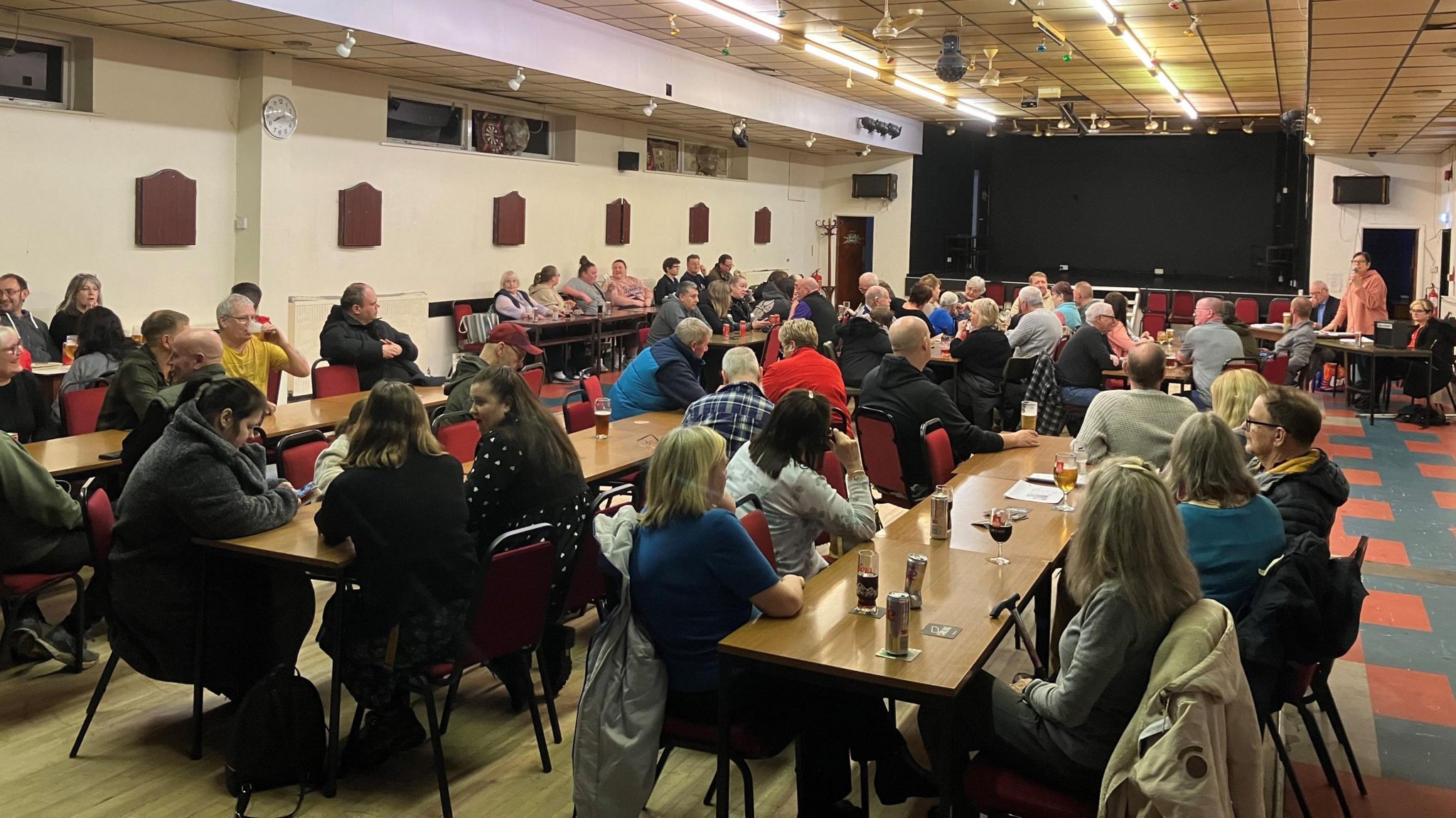 A picture of a public meeting, people sitting at long tables in a function room
