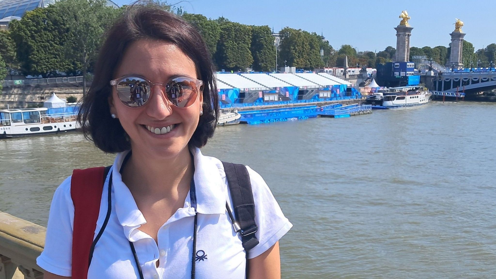 A young woman with dark hair in a bob, wearing a white polo shirt and a rucksack, stands on a bridge over a river