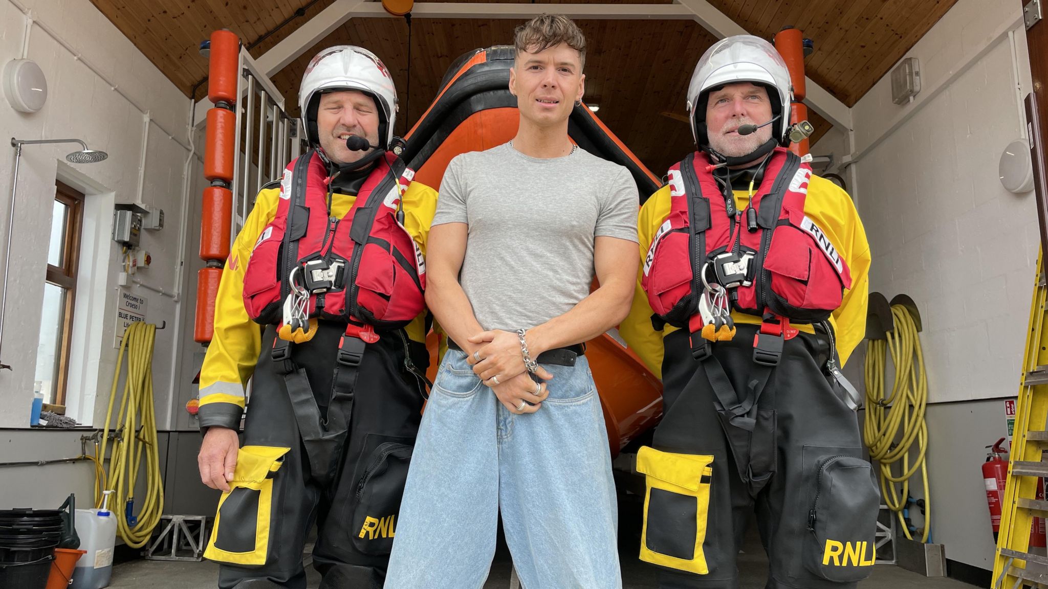 Ren Gill posiert zwischen zwei voll ausgerüsteten Mitgliedern der RNLI-Besatzung in der Rettungsbootstation Beaumaris auf Anglesey