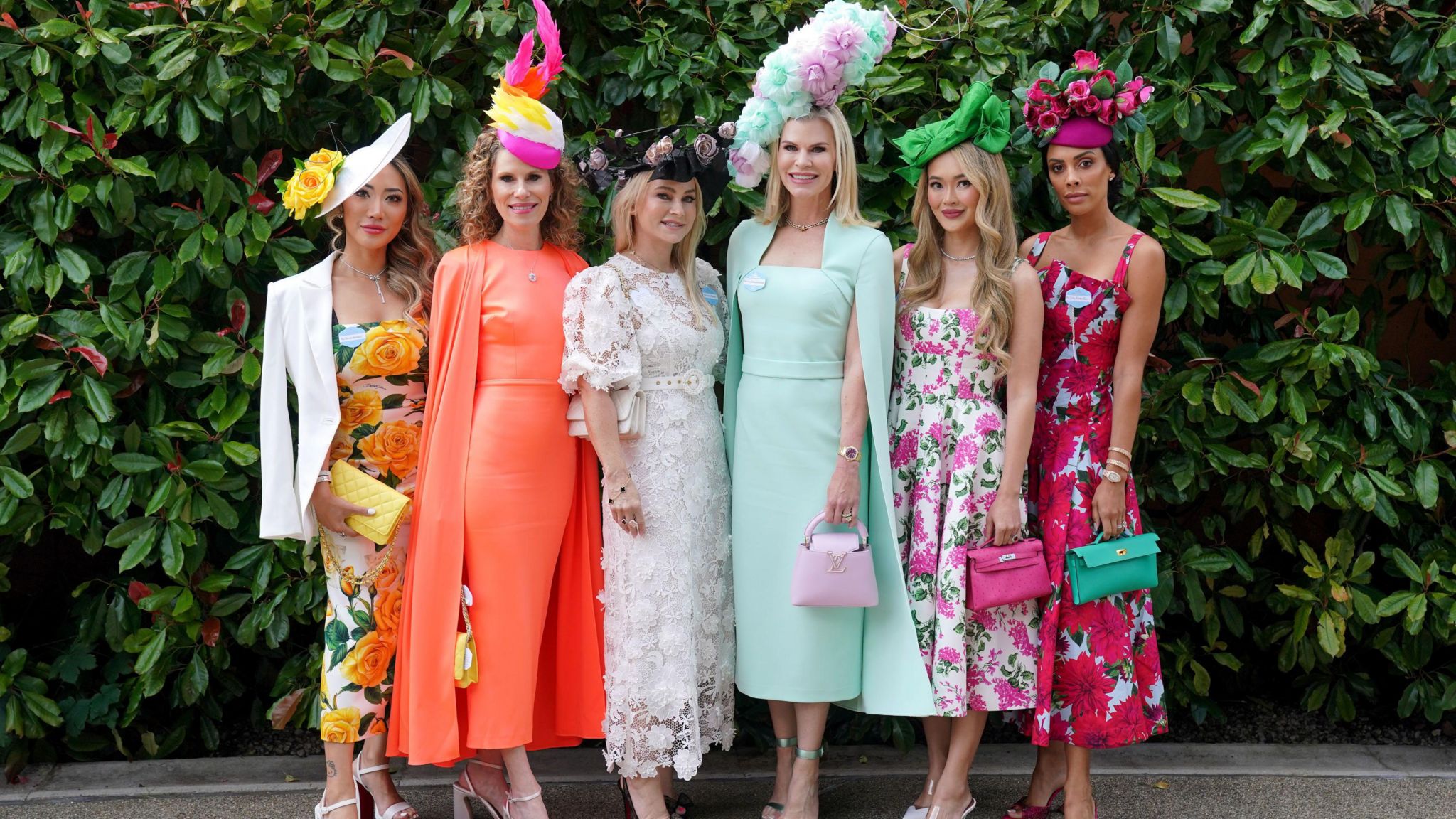 Six young women standing in front of a large green bush, dressed in smart summer dresses and extravagant hats