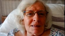 Annette Smith lying on a bed, with white hair and glasses
