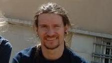 Mark Kennedy wearing a t-shirt and smiling with a building behind him