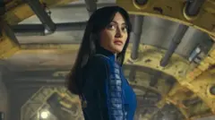 A woman with long black hair stands in the middle of a steampunk-like room. There are pipes and girders visible with yellow and grey colours. The woman has wide eyes and is wearing a deep blue jumpsuit.
