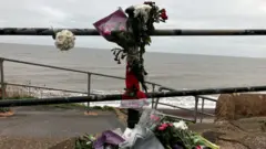 BBC Flowers have been placed near a beach following the tragedy on Christmas Day. The flowers have been placed next to a metal railing. The sea is lapping on the shoreline in the background. A note on one of the bouquets says 'rest in peace'.