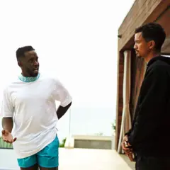 Sean Combs (left) and Phil Pines (right) talk outdoors near a building with an ocean view in the background; Combs wears a white t-shirt and blue shorts, Pines is in a black hoodie.