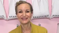 Marcia Kilgore with cropped short brown hair smiling at the camera with a blurred background wearing a nude coloured round neck top