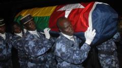 Togolese gendarmes carry di coffin of one of di victims of di attack, wrapped in di national flag