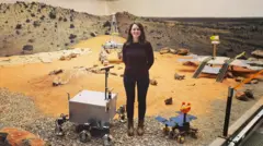 A woman wearing a brown jumper and black trousers stands on a simulated surface of Mars with orange sand and stones, with prototypes at her feet