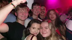 A crowded group photograph taken in a nightclub or live music venue. Several people are closely gathered together and filling most of the frame. Two people are positioned in the foreground, standing cheek-to-cheek near the centre, while additional people stand behind them in rows extending into the background. On the left side of the image, a raised arm wearing a multicoloured beaded bracelet reaches upward and partially enters the frame. The room is packed with people, with only small glimpses of walls and fixtures visible. Bright pink and purple club lighting shines from the upper right corner and reflects across faces, hair and clothing throughout the crowd. The setting appears busy and energetic, with people standing shoulder to shoulder in a lively indoor nightlife environment.
