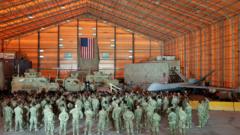 Mike Pence no parlatório dentro de base, dentro da qual se vê bandeira americana, tanques e avião, assistido por dezenas de soldados