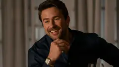 Lionsgate Actor Brandon Sklenar appearing in film The Housemaid. He is smiling whilst holding his chin with his hand. He is wearing a blue shirt.
