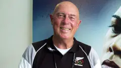 Getty Images Les Mills, a man with bushy greying eyebrows and a bald head who is smiling straight ahead - he is wearing a black, white and grey sports top which has the Olympic rings, a silver fern logo and the words 'New Zealand' on the right-hand-side - he is standing in front of a poster, on which part of a woman's face can be seen 
