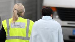 A woman in a hi-vis UK Visas and Immigration jacket walks beside a man wrapped in a towel