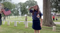 A young girl scout stands next to a tree saluting.